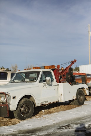 A vintage tow truck with a white cab and a rusty red towing mechanism is parked on a patch of grass and snow. The truck appears weathered, with visible rust and worn paint. In the background, other vehicles and a utility pole can be seen under a clear blue sky.
