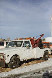 A vintage tow truck with a white cab and a rusty red towing mechanism is parked on a patch of grass and snow. The truck appears weathered, with visible rust and worn paint. In the background, other vehicles and a utility pole can be seen under a clear blue sky.