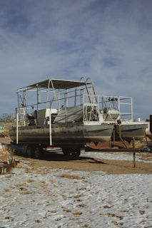 A large pontoon boat is positioned on a trailer, situated on a snow-covered ground. The boat features metal framework and protective covering, with visible equipment and components onboard. In the background, a few structures and leafless trees are partially visible under a sky with a mix of clouds.