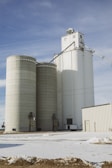 Several large cylindrical silos made of concrete stand prominently against a partly cloudy blue sky. The silos are connected to a white building on the right, with snow covering the ground in the foreground.