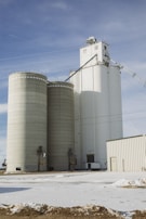 Several large cylindrical silos made of concrete stand prominently against a partly cloudy blue sky. The silos are connected to a white building on the right, with snow covering the ground in the foreground.