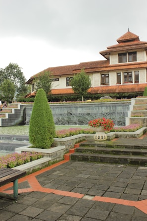 A landscaped garden area with tiered water features and a prominent, well-trimmed evergreen shrub. The garden is surrounded by stone pathways and staircases with decorative flower beds, including a large pot of red flowers. In the background is a large building with a red-tiled roof and multiple windows, resembling a traditional or colonial-style architecture.