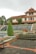 A landscaped garden area with tiered water features and a prominent, well-trimmed evergreen shrub. The garden is surrounded by stone pathways and staircases with decorative flower beds, including a large pot of red flowers. In the background is a large building with a red-tiled roof and multiple windows, resembling a traditional or colonial-style architecture.