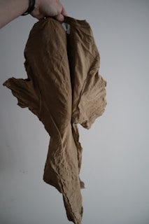 A crumpled beige cloth or garment is held up by a hand and appears to have a casual, loose fit. The fabric shows visible wrinkles and texture. The background is plain and neutral, emphasizing the texture and color of the fabric.