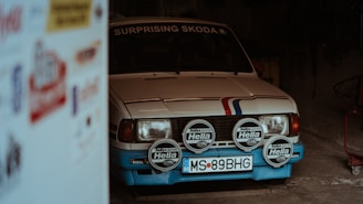A vintage car with a white body and blue accents is parked partially inside a dimly lit garage. It has a rally number plate and multiple Hella spotlights attached to the front. The word 'Surprising' is written on its windshield. The side of the image shows blurred advertisements.