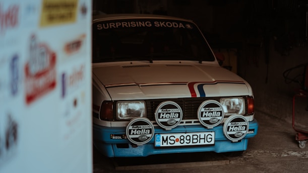 A vintage car with a white body and blue accents is parked partially inside a dimly lit garage. It has a rally number plate and multiple Hella spotlights attached to the front. The word 'Surprising' is written on its windshield. The side of the image shows blurred advertisements.