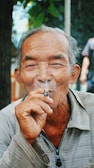 An elderly man with gray hair and a gentle expression is enjoying a cigarette. He wears a light-colored jacket with a zippered front, sitting comfortably outdoors with foliage in the background. His eyes are slightly closed, conveying a sense of relaxation.