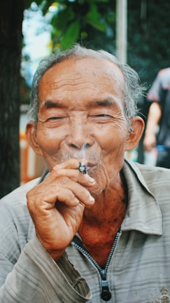 An elderly man with gray hair and a gentle expression is enjoying a cigarette. He wears a light-colored jacket with a zippered front, sitting comfortably outdoors with foliage in the background. His eyes are slightly closed, conveying a sense of relaxation.