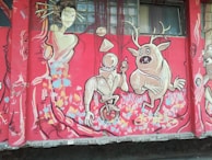 A colorful mural featuring abstract and cartoonish characters. A geometric-headed figure on a unicycle is holding a lollipop, while a quirky creature with antlers strikes a playful pose beside it. A stylized feminine figure in an elegant dress with flowing hair appears on the left, surrounded by vibrant patterns and shapes. The background is predominantly red with splashes of blue, pink, and yellow.