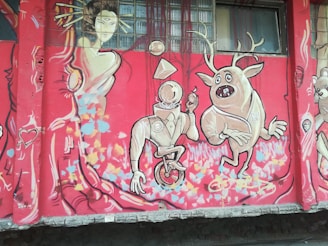 A colorful mural featuring abstract and cartoonish characters. A geometric-headed figure on a unicycle is holding a lollipop, while a quirky creature with antlers strikes a playful pose beside it. A stylized feminine figure in an elegant dress with flowing hair appears on the left, surrounded by vibrant patterns and shapes. The background is predominantly red with splashes of blue, pink, and yellow.