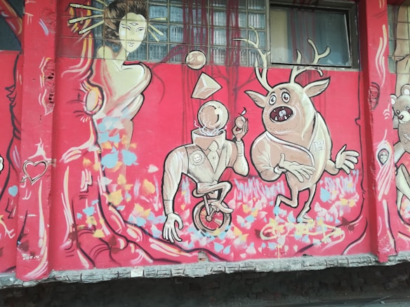 A colorful mural featuring abstract and cartoonish characters. A geometric-headed figure on a unicycle is holding a lollipop, while a quirky creature with antlers strikes a playful pose beside it. A stylized feminine figure in an elegant dress with flowing hair appears on the left, surrounded by vibrant patterns and shapes. The background is predominantly red with splashes of blue, pink, and yellow.