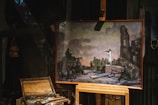A painting on an easel depicts a dramatic scene with several figures in military attire gathered around a person standing on a platform. The background features ruins and a cloudy sky. Below the easel, there is an artist's box with paintbrushes and other painting materials.