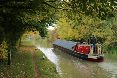 A narrowboat travels along a serene canal, surrounded by lush green trees on both sides. The canal is calm and reflects the canopy of branches and foliage above. A person stands at the back of the narrowboat, steering it gently along the waterway. The path along the canal is bordered with grass and scattered autumn leaves.