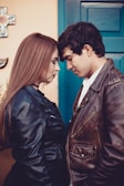 A young couple stands close together, facing each other with their foreheads touching, creating an intimate and affectionate pose. Both are wearing leather jackets; the woman has long hair, and the man has short, dark hair. The background shows a colorful wall with a cross and a blue door.