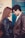 A young couple stands close together, facing each other with their foreheads touching, creating an intimate and affectionate pose. Both are wearing leather jackets; the woman has long hair, and the man has short, dark hair. The background shows a colorful wall with a cross and a blue door.