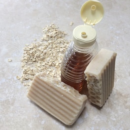A clear plastic bottle filled with amber-colored liquid is positioned next to two bars of textured beige soap. Oats are scattered across the light stone surface, providing a rustic and natural setting.