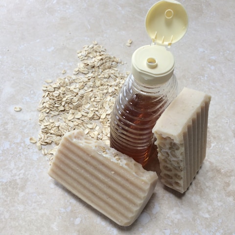 A clear plastic bottle filled with amber-colored liquid is positioned next to two bars of textured beige soap. Oats are scattered across the light stone surface, providing a rustic and natural setting.