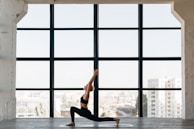 A person practices yoga indoors, performing a lunge pose on a mat in front of a large window. The room has a spacious and open feel, with city buildings visible in the background through the window.