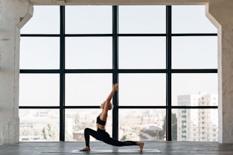 A person practices yoga indoors, performing a lunge pose on a mat in front of a large window. The room has a spacious and open feel, with city buildings visible in the background through the window.
