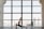 A person practices yoga indoors, performing a lunge pose on a mat in front of a large window. The room has a spacious and open feel, with city buildings visible in the background through the window.