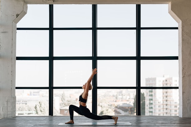 A person practices yoga indoors, performing a lunge pose on a mat in front of a large window. The room has a spacious and open feel, with city buildings visible in the background through the window.