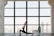 A person practices yoga indoors, performing a lunge pose on a mat in front of a large window. The room has a spacious and open feel, with city buildings visible in the background through the window.
