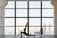 A person practices yoga indoors, performing a lunge pose on a mat in front of a large window. The room has a spacious and open feel, with city buildings visible in the background through the window.
