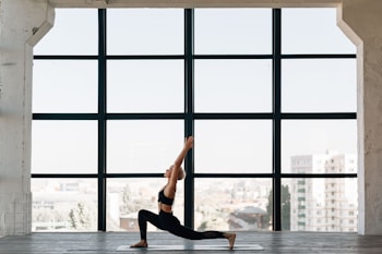 A person practices yoga indoors, performing a lunge pose on a mat in front of a large window. The room has a spacious and open feel, with city buildings visible in the background through the window.