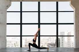 A person practices yoga indoors, performing a lunge pose on a mat in front of a large window. The room has a spacious and open feel, with city buildings visible in the background through the window.