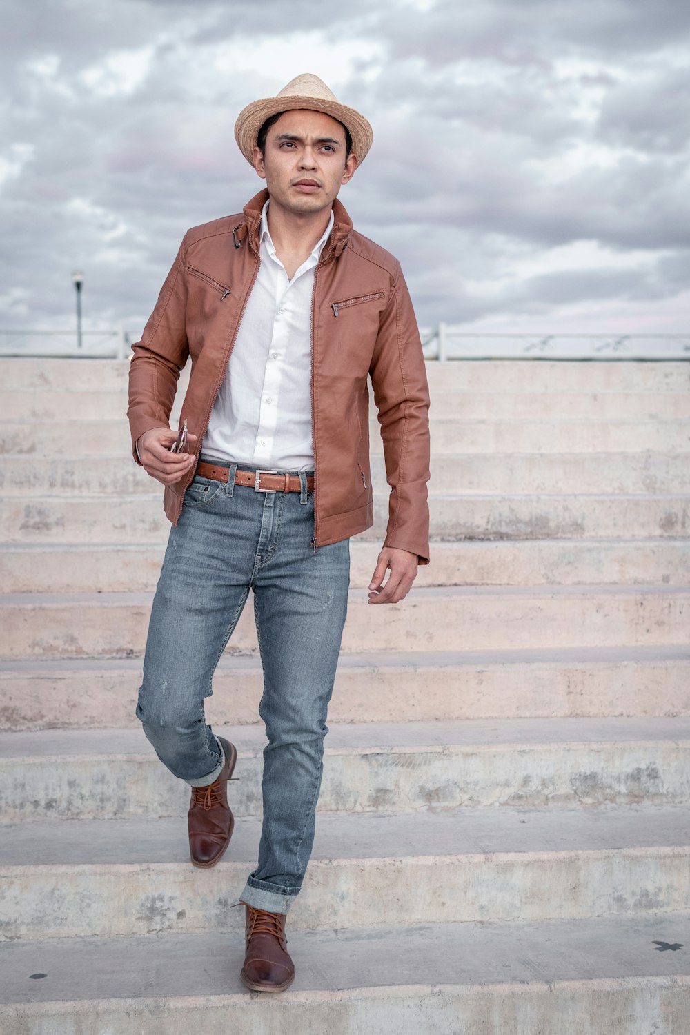 uomo in giacca di pelle marrone e jeans blu denim in piedi sulle scale di cemento bianco durante il giorno
