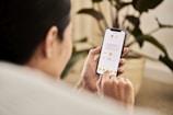 A person holding a smartphone and interacting with a mobile app that displays a document. The individual is sitting indoors, likely in a relaxed setting with a houseplant visible in the background.