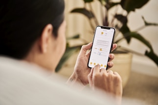 A person holding a smartphone and interacting with a mobile app that displays a document. The individual is sitting indoors, likely in a relaxed setting with a houseplant visible in the background.