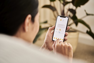 A person holding a smartphone and interacting with a mobile app that displays a document. The individual is sitting indoors, likely in a relaxed setting with a houseplant visible in the background.