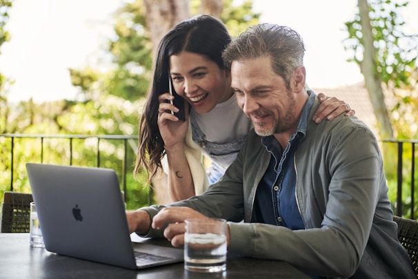 A man and woman are sitting outside on a patio, engaged with a laptop. The woman is holding a smartphone to her ear while smiling, and the man is focused on the laptop screen. They both appear to be in a cheerful and collaborative mood. The background shows lush greenery, suggesting they are in a garden or outdoor setting.