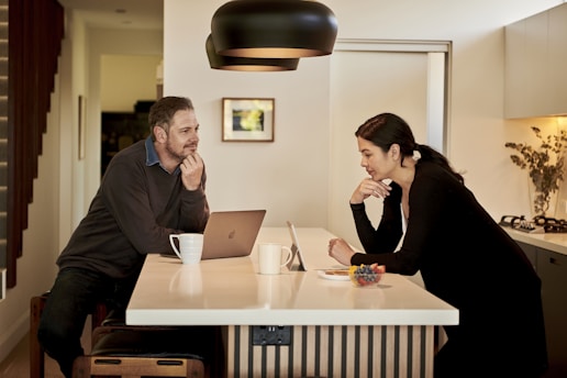 Two people are engaged in conversation at a kitchen counter, each with a laptop. A man sits on a stool, while a woman stands, both appearing focused on their discussion. The kitchen features a modern design with a large hanging light fixture and a vase with flowers in the background. Mugs and a small bowl of fruit are on the counter, adding a casual touch to the scene.