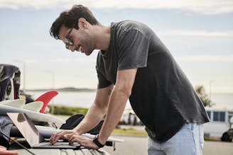 A man wearing sunglasses is leaning over a laptop on the back of a vehicle. Several surfboards with colorful fins are visible next to him. The scene takes place outdoors near a beach, and the background includes a blurred view of the ocean and some distant hills.