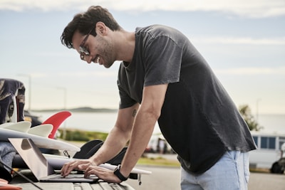 A man wearing sunglasses is leaning over a laptop on the back of a vehicle. Several surfboards with colorful fins are visible next to him. The scene takes place outdoors near a beach, and the background includes a blurred view of the ocean and some distant hills.