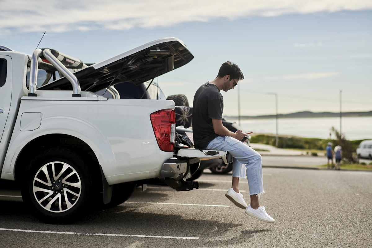 A man sits on the lowered tailgate of a white pickup truck, reading his phone in a seaside parking lot
