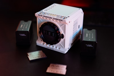 A white, cube-shaped RED Digital Cinema camera sits prominently on a dark surface, surrounded by two camera batteries and memory cards. The scene is dimly lit with a red hue in the background, providing a cinematic and professional ambiance.