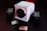 A white, cube-shaped RED Digital Cinema camera sits prominently on a dark surface, surrounded by two camera batteries and memory cards. The scene is dimly lit with a red hue in the background, providing a cinematic and professional ambiance.