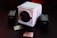 A white, cube-shaped RED Digital Cinema camera sits prominently on a dark surface, surrounded by two camera batteries and memory cards. The scene is dimly lit with a red hue in the background, providing a cinematic and professional ambiance.