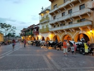 A vibrant street scene with historic colonial-style buildings featuring balconies and arches. The street is lined with horse-drawn carriages and people are strolling, engaging in leisurely activities. The setting appears lively, with a warm and inviting ambiance enhanced by the colors of the buildings and evening lights.