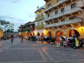 A vibrant street scene with historic colonial-style buildings featuring balconies and arches. The street is lined with horse-drawn carriages and people are strolling, engaging in leisurely activities. The setting appears lively, with a warm and inviting ambiance enhanced by the colors of the buildings and evening lights.