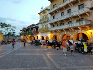 A vibrant street scene with historic colonial-style buildings featuring balconies and arches. The street is lined with horse-drawn carriages and people are strolling, engaging in leisurely activities. The setting appears lively, with a warm and inviting ambiance enhanced by the colors of the buildings and evening lights.