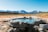 A small natural hot spring surrounded by rocks, situated in a dry, rocky landscape. The water is a tranquil blue and emits a light steam, suggesting warmth. Snow-capped mountains are visible in the background under a clear blue sky, adding contrast to the arid foreground.
