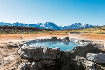 A small natural hot spring surrounded by rocks, situated in a dry, rocky landscape. The water is a tranquil blue and emits a light steam, suggesting warmth. Snow-capped mountains are visible in the background under a clear blue sky, adding contrast to the arid foreground.