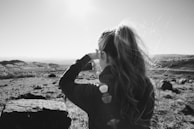 A black and white image capturing a person with long hair tied in a ponytail. They are wearing a jacket, facing away from the camera, and appear to be photographing or looking out over a vast landscape. The scene is sunlit, creating lens flares and a serene atmosphere.