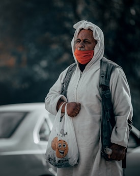 An elderly person wearing a white hooded garment and a face mask holds a plastic bag with a cartoon face printed on it. The background shows an out-of-focus car.