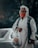 An elderly person wearing a white hooded garment and a face mask holds a plastic bag with a cartoon face printed on it. The background shows an out-of-focus car.