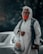 An elderly person wearing a white hooded garment and a face mask holds a plastic bag with a cartoon face printed on it. The background shows an out-of-focus car.